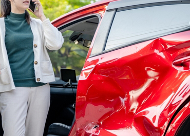 Woman standing by her severely damaged red car, talking on a phone. Title: The Benefits Of Hiring A Car Accident Lawyer.